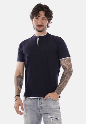 Uomo con capelli ricci che indossa una maglia henley blu navy a maniche corte e jeans grigi chiari strappati, mostrando tatuaggi su entrambe le braccia.