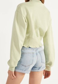 Kvinna i ljusgrön långärmad topp med smockad midja och ljusblå denimshorts med rullad nederkant, sedd bakifrån.