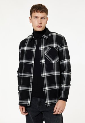 Ung mand iført sort og hvid ternet knap-op skjorte over sort rullemaskine, stående mod en ensfarvet hvid baggrund.