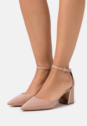 Beige ankle strap heels with a pointed toe and block heel featuring a metal accent. Smooth leather finish with a clean, sleek design.
