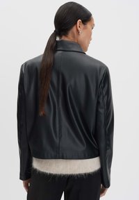 Giacca di pelle nera con colletto sartoriale, texture liscia e vestibilità corta, indossata sopra un maglione in maglia di colore chiaro.