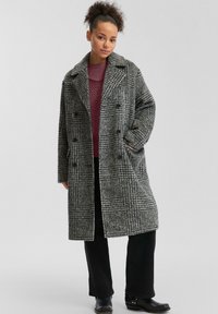 Manteau en mélange de laine à carreaux en gris et noir, présentant un design à double boutonnage, de grands revers et deux poches à l'avant. Pantalon noir et bottes visibles.