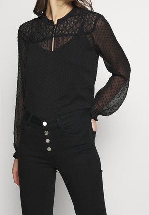 Kobieta ubrana w prześwitującą czarną bluzkę z długim rękawem w groszki oraz czarne, wysokie spodnie z czterema srebrnymi guzikami.