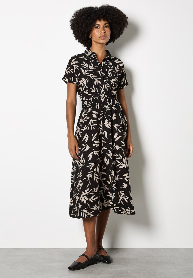 Schwarzes Kleid mit kurzen Ärmeln, ausgestattet mit cremefarbenen Blattmustern, einem Knopfkragen und einem elastischen Taillenzug; kombiniert mit schwarzen Ballerinas.