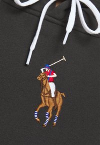 Bestickter Polo-Spieler in rotem Shirt und blauem Helm auf braunem Pferd mit blau geringelten Beinschützern auf dunklem Stoff neben weißen Kapuzenzugschnüren.