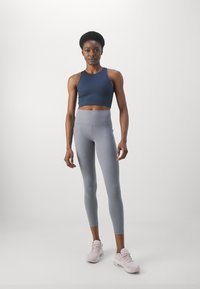 Ναυτικό κορμάκι, ψηλόμεσα γκρι κολάν και ανοιχτό ροζ αθλητικά παπούτσια. Το μοντέλο στέκεται μπροστά σε λευκό απλό φόντο.