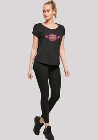 Melna īsām piedurknēm T-kreklis ar Wonder Woman grafiku, savienojumā ar melniem legingiem un melniem sporta apaviem, uz gaiši pelēka fona.
