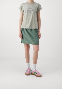 Grå bomullströja med rullade ärmar och logotyp, tillsammans med en grön kjol med sidoknytningar och rosa mockaskor med gröna ränder.