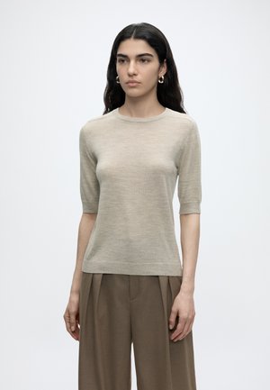 Žena s dlhými tmavými vlasmi, ktorá má na sebe zlaté kruhové náušnice, béžový krátky pletený top a voľné hnedé nohavice, stojaca pred jednoduchým svetlým pozadím.