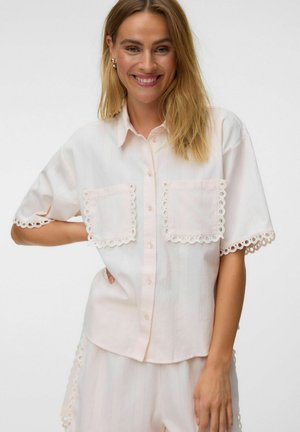 Femme souriante aux cheveux blonds porte une chemise ample rose clair et un short assorti avec un bord en dentelle œillets sur les poches, les manches et les côtés.