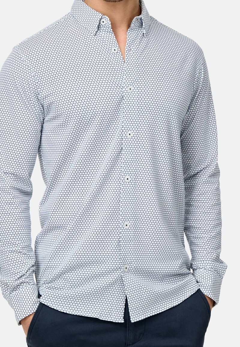 Bílá košile s drobným námořnicky modrým geometrickým vzorem, límcem na knoflíky, dlouhými rukávy a rovnou lemovkou. Vyrobená z lehké látky.