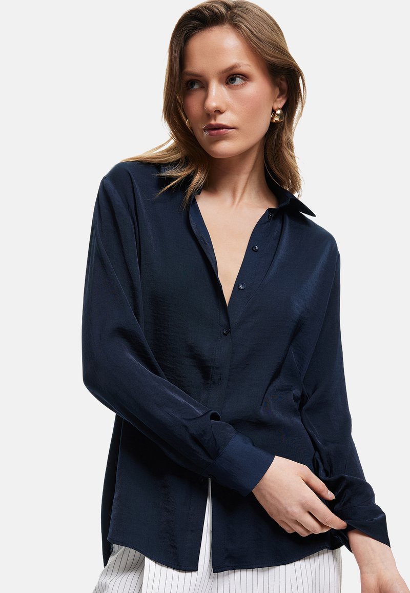 Navy blue button-up shirt made of smooth fabric, featuring a collar, long sleeves, and a relaxed fit with a slightly curved hem.