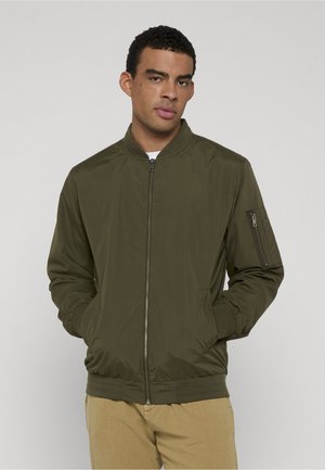 Ung mann med kort krøllete hår iført en olivengrønn bomberjakke med glidelås og beige bukser, hendene i lommene, stående mot en grå bakgrunn.