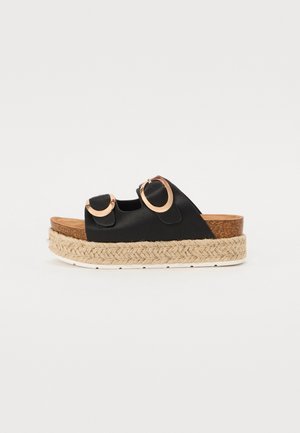 Sort platform slidesandal med to store guldspænder og flettet jutesål på hvid baggrund.