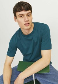 Turquoise t-shirt met korte mouwen en ronde hals, gecombineerd met blauwe denim jeans. Het model zit, met de armen rustend op een groen object.