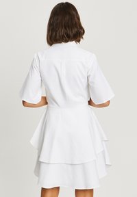 Robe blanche à manches courtes avec un ourlet à volants en cascade, un col et un dos boutonné. Le tissu semble lisse et est léger.