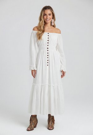 Vrouw in een lange witte off-shoulder jurk met knopen aan de voorkant en bruine open-toe enkellaarzen met franjes.
