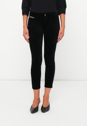 Femme portant un pantalon noir slim cropped avec un détail de poche ornée de bijoux et des talons noirs pointus, debout sur fond blanc.