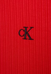 Röd tyg med vertikal ribbad textur som har en svart broderad "cK"-logotyp i mitten.