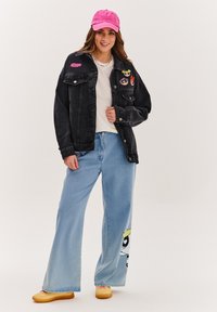 Czarna dżinsowa kurtka z przednimi kieszeniami i kolorowymi łatami, zestawiona z jasnoniebieskimi spodniami szerokimi w nogawkach z nadrukiem z kreskówki oraz różową czapką.