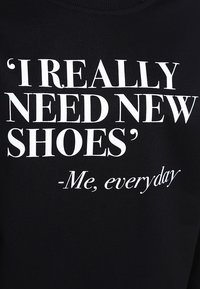 El texto sobre la tela negra dice: "'REALMENTE NECESITO ZAPATOS NUEVOS' -Yo, todos los días" en letras mayúsculas blancas y cursivas.