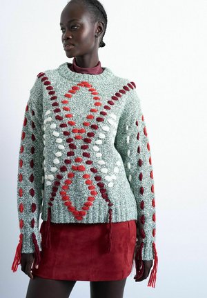 Donna che indossa un maglione lavorato a maglia grossa con motivi in rilievo rossi, bianchi e marroni abbinato a una minigonna in camoscio rossa.