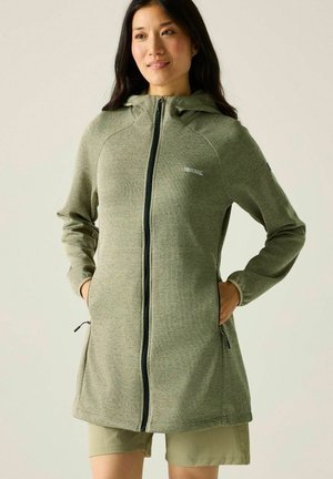 Femme portant une veste à capuche zippée vert olive, les mains dans les poches, debout sur un fond clair.