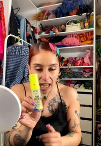 Femme avec des barrettes colorées et des tatouages tenant une bouteille jaune de soin pour la peau NYX Plump Right Back devant une étagère encombrée de vêtements et d'accessoires.