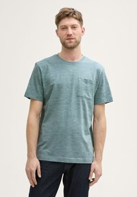 Camiseta de manga corta en color azul verdoso, hecha de tela texturizada, con un bolsillo en el pecho y un cuello redondo, combinada con jeans de mezclilla oscura.
