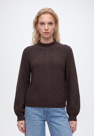 Femme aux cheveux blonds portant un pull en maille brun foncé avec des motifs torsadés et un jean bleu clair, debout devant un fond clair uni.