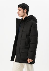 Manteau rembourré noir avec capuche, présentant une finition texturée, des poches avant et une fermeture éclair. Design mi-long, isolation légère.