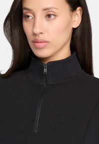 Svart zip-up sweatshirt med hög krage, ribbad krage, mjukt material och en liten, diskret logotyp på bröstet.