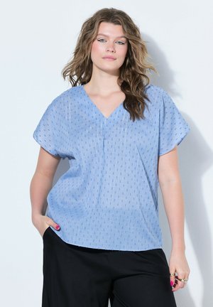 Kvinde med bølget hår iført en lyseblå mønstret V-hals bluse og sorte bukser, stående med den ene hånd i lommen mod en hvid baggrund.