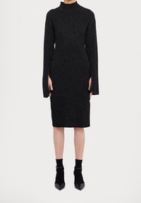 Femme portant une robe en maille côtelée noire ajustée avec des manches longues fendues aux poignets, associée à des bottines noires.