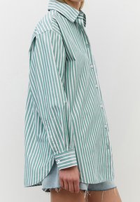 Camicia a righe verde e bianca, con colletto classico, maniche lunghe, chiusura con bottoni frontale e vestibilità ampia con orlo arrotondato.