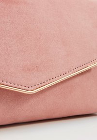 Close-up of a pink suede clutch bag with a gold metal trim along the flap edge on a white surface.