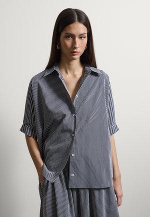 Joven mujer con cabello largo y oscuro, que lleva una camisa suelta de manga corta con rayas verticales blancas y negras y una falda a juego sobre un fondo liso.