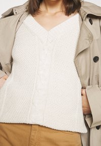 Sweter typu V-neck w kremowym kolorze z wzorem w warkocze. Noszony pod beżowym płaszczem trench, z teksturowanym materiałem i luźnym krojem.
