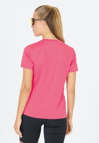 Lyserød træningsshirt med korte ærmer, lavet af letvægtsstof, med syede skuldersømme. Båret med sorte shorts.