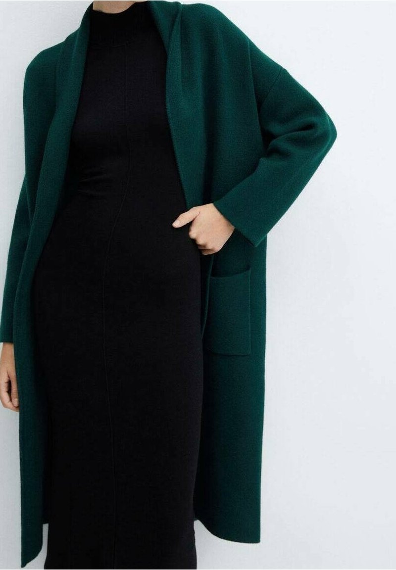 Femme portant un long manteau vert foncé par-dessus une robe noire ajustée, avec une main dans la poche, sur un fond blanc uni.