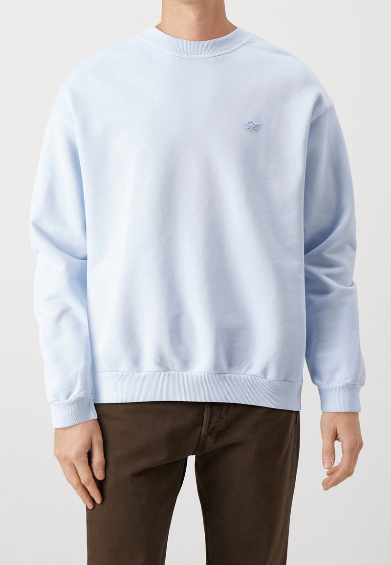 Homme portant un sweat-shirt à col rond à manches longues bleu clair avec un petit logo sur la poitrine et un pantalon marron foncé, debout les mains détendues.
