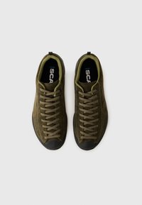 Chaussures vert olive foncé avec une tige en daim, des lacets plats et un embout en caoutchouc. L'intérieur est doublé d'un vert plus clair avec un logo sur la semelle intérieure.