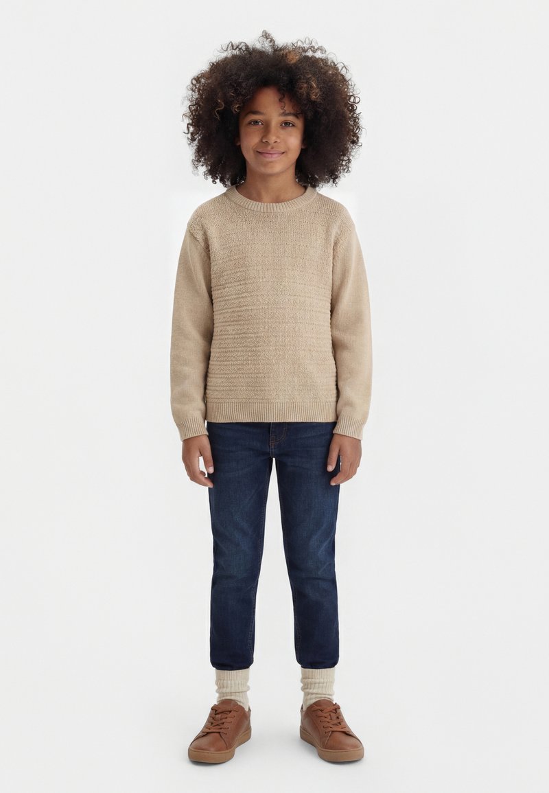 Bambino con capelli ricci che indossa un maglione beige lavorato a maglia, jeans blu scuro, calzini beige e scarpe da ginnastica marroni, in piedi rivolto verso l'obiettivo.