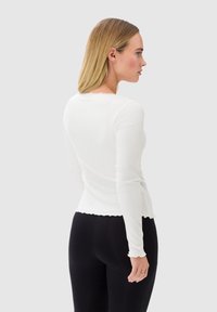 Biała bluzka z długim rękawem o teksturowanym, prążkowanym wzorze, z falbaniastym dekoltem i dopasowanym krojem, w zestawieniu z czarnymi legginsami.