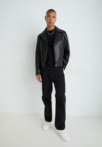 Must musta jalgratturi jakk, millel on tõmblukuga taskud, kantud musta graafilise kapuutsiga särk üle, koos mustade teksade ja valgete tossudega.