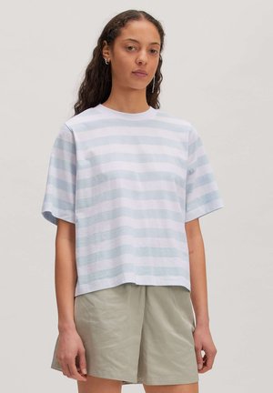 Jonge vrouw met lang krullend haar draagt een lichtblauw-wit gestreept T-shirt met korte mouwen en beige short, staand tegen een effen achtergrond.