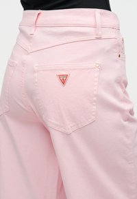 Ροζ τζιν παντελόνι με εφαρμοστή μέση, δύο πίσω τσέπες και ένα μικρό κόκκινο τριγωνικό λογότυπο στην δεξιά τσέπη. Λείο υλικό, casual σχεδιασμός.