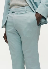 Άνδρας μοντέλο φορώντας ανοιχτό μπλε παντελόνι κοστουμιού και ταιριαστό σακάκι, με το δεξί χέρι στην τσέπη, μερικώς ορατό μπεζ πλεκτό τοπ.