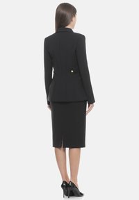 Blazer negro entallado con cintura peplum y abertura trasera, detallado con botones de tono dorado. Combinado con una falda lápiz ajustada a juego.