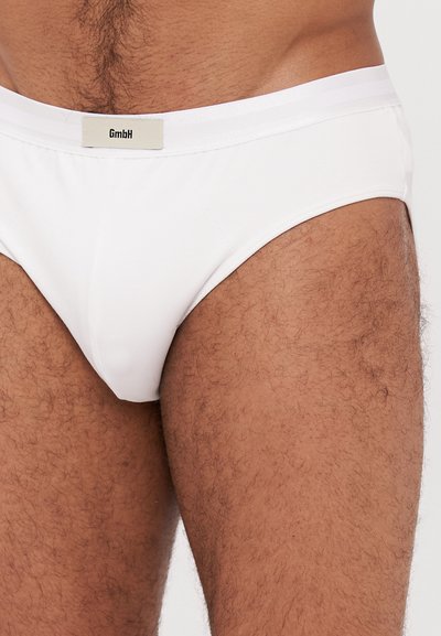 Culottes blanches en tissu extensible, dotées d'une fine ceinture élastique avec une étiquette. Texture douce et design ajusté.